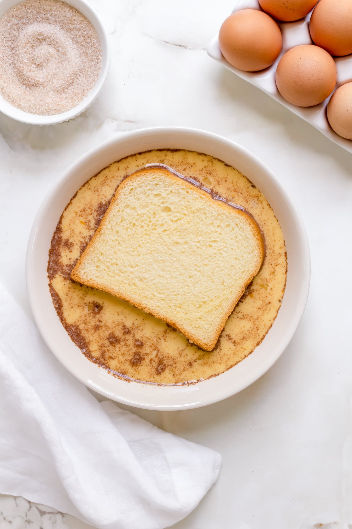 brioche soaking in white bowl of custard with container of eggs and bowl of cinnamon and sugar. 