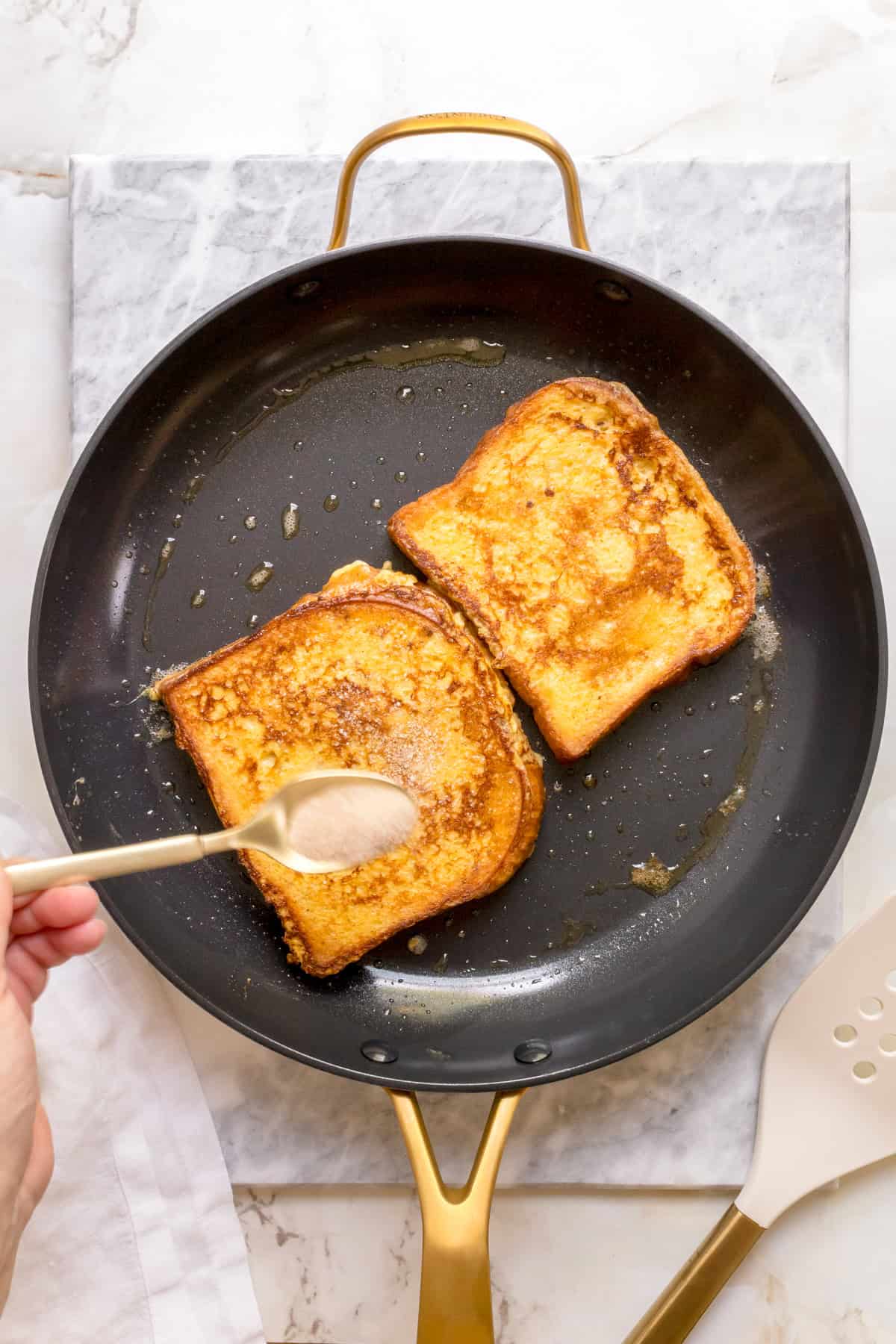 gold spoon sprinkling cinnamon and sugar over brioche French toast in buttered frying pan.