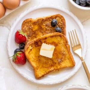 two slices of brioche French toast on white plate with gold fork, berries and eggs.