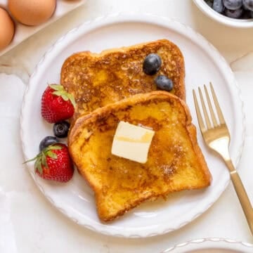 two slices of brioche French toast on white plate with gold fork, berries and eggs.