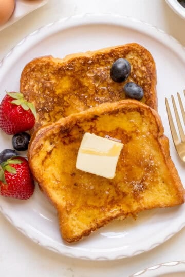 two slices of brioche French toast on white plate with gold fork, berries and eggs.