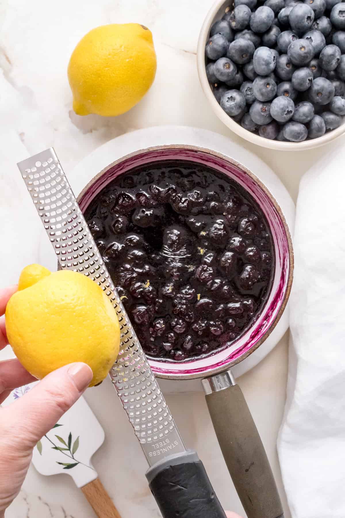 zesting fresh lemon into blueberry compote in white saucepan.