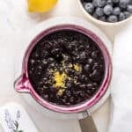 blueberry compote in white saucepan with a bowl of blueberries and a lemon.