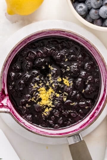blueberry compote in white saucepan with a bowl of blueberries and a lemon.