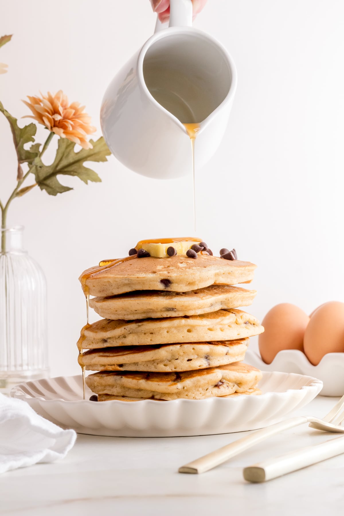 pouring maple syrup over chocolate chip pancakes on white plate with gold fork.