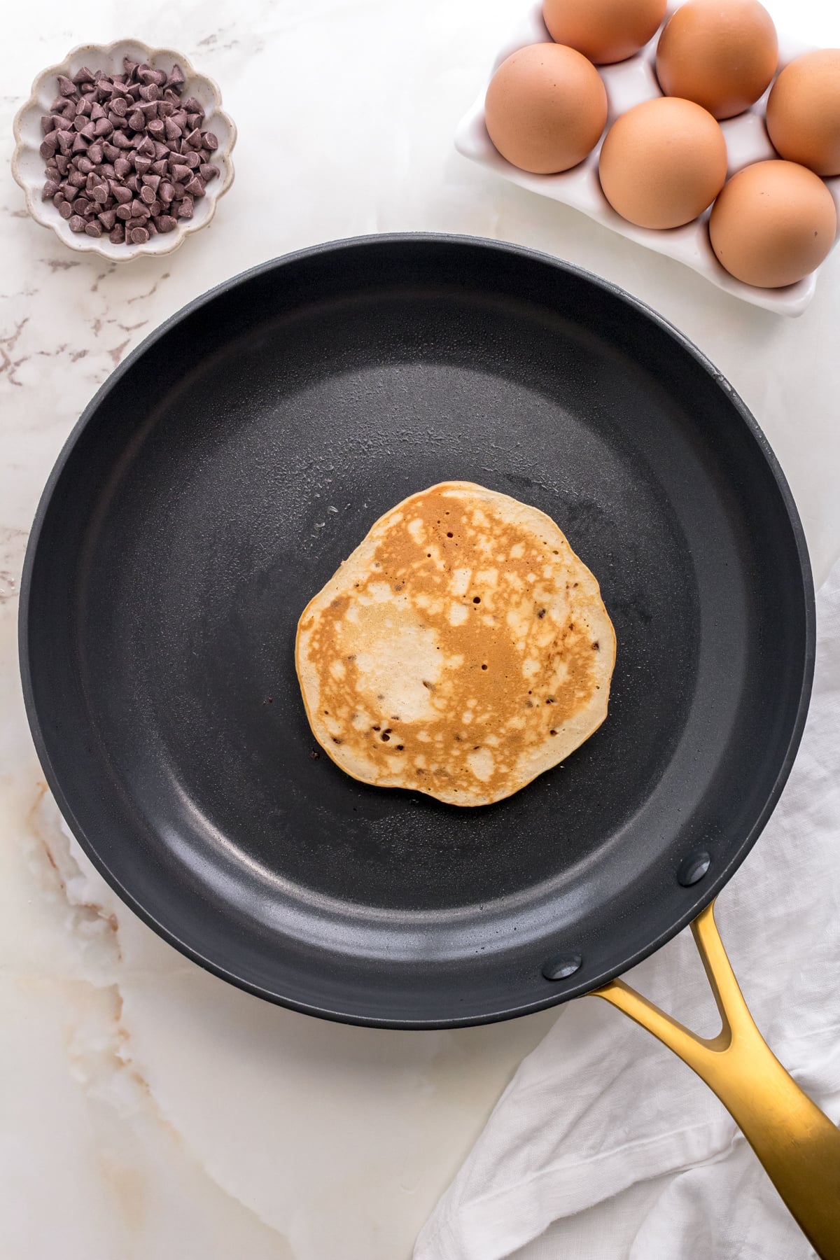 chocolate chip pancake cooking in gold frying pan with eggs and bowl of chocolate chips.