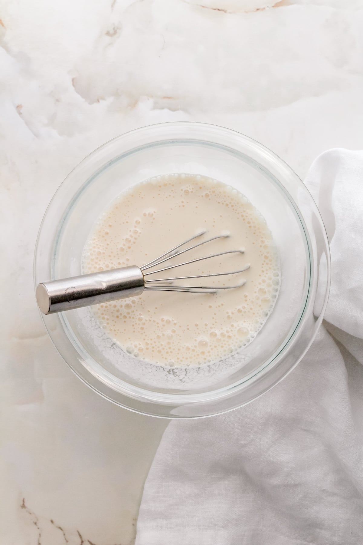 small glass mixing bowl with milk and sourdough discard on white background with a whisk.