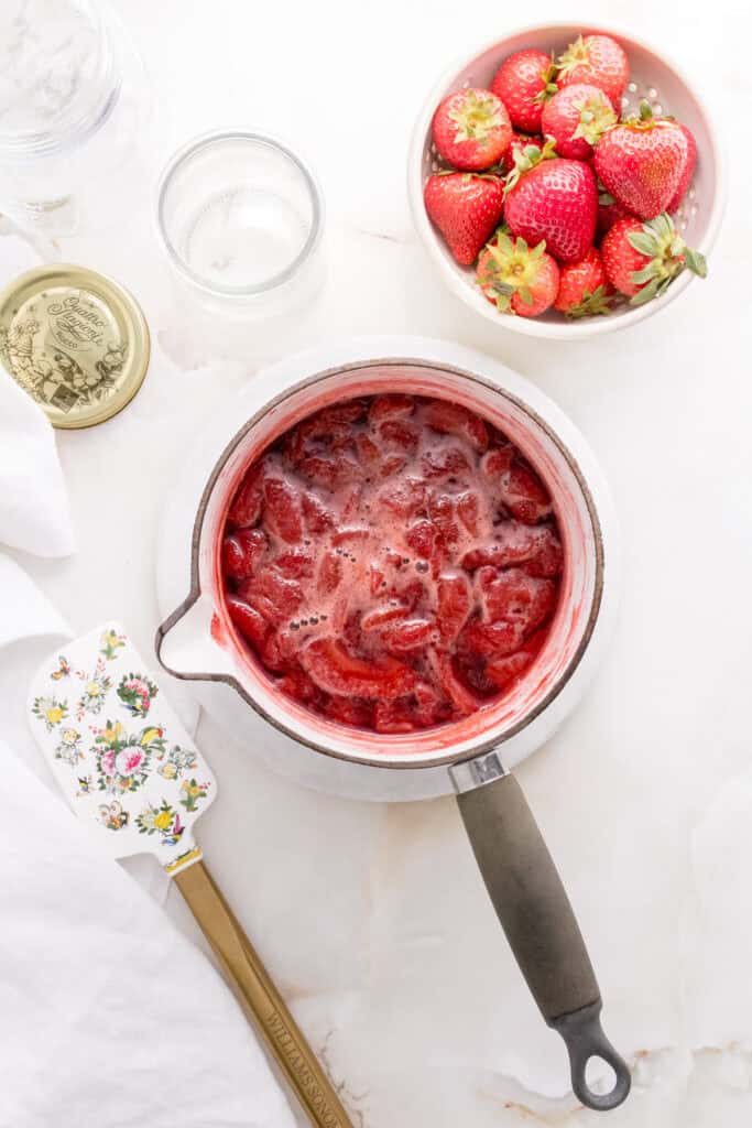 strawberry compote simmering in white saucepan with spatula and bowl of strawberries.