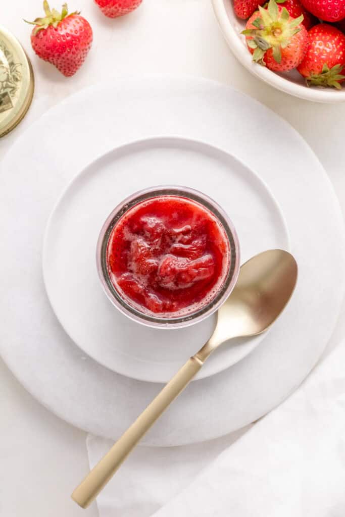 strawberry compote in a small glass jar with a bowl of strawberries and a gold spoon.