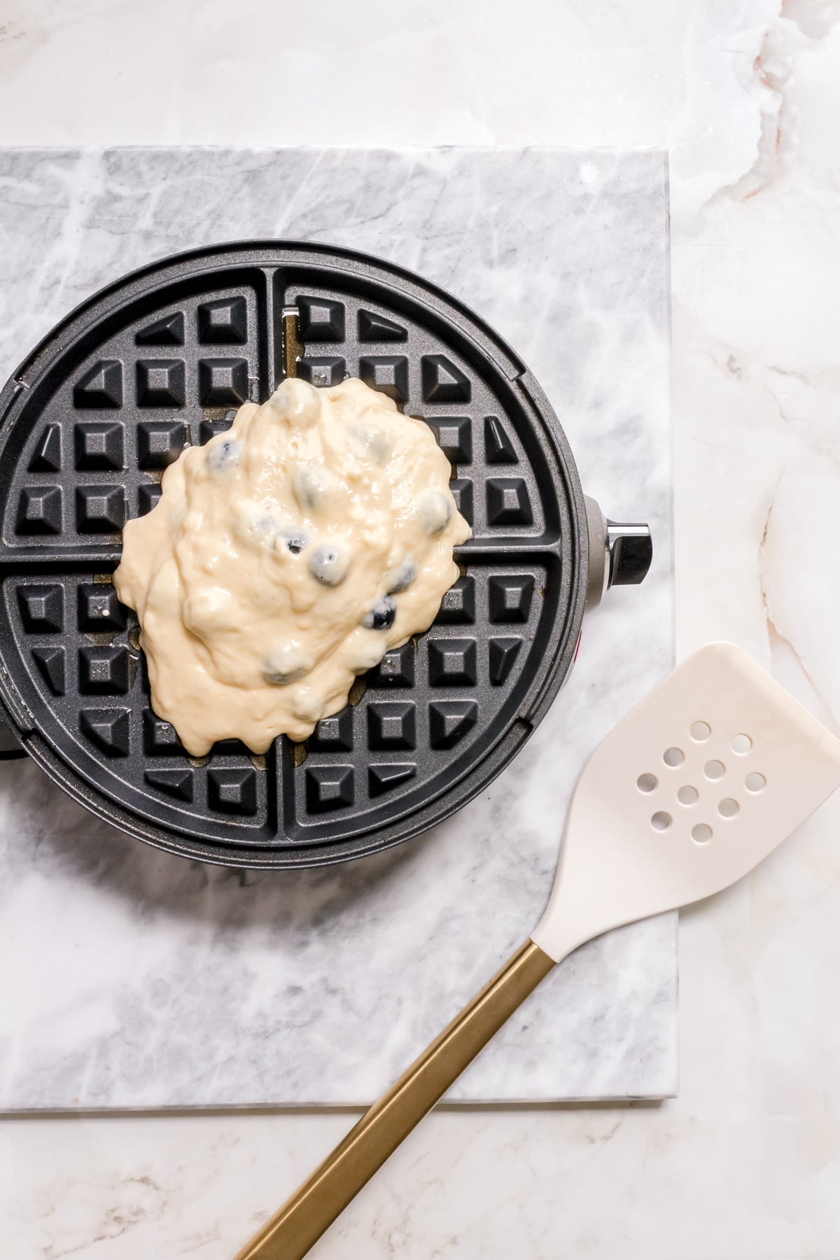 waffle iron with blueberry waffle batter and white and gold spatula.