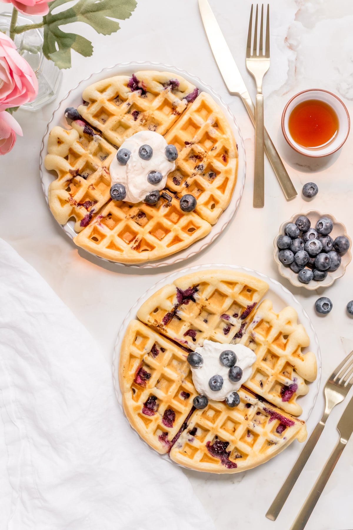 two white plates of blueberry waffles with whipped cream and maple syrup.