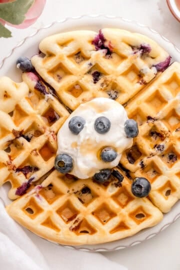 blueberry waffles with whipped cream on white plate with gold fork and knife.