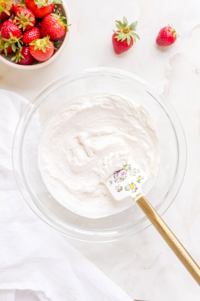 homemade whipped cream in glass bowl with spatula and bowl of strawberries.