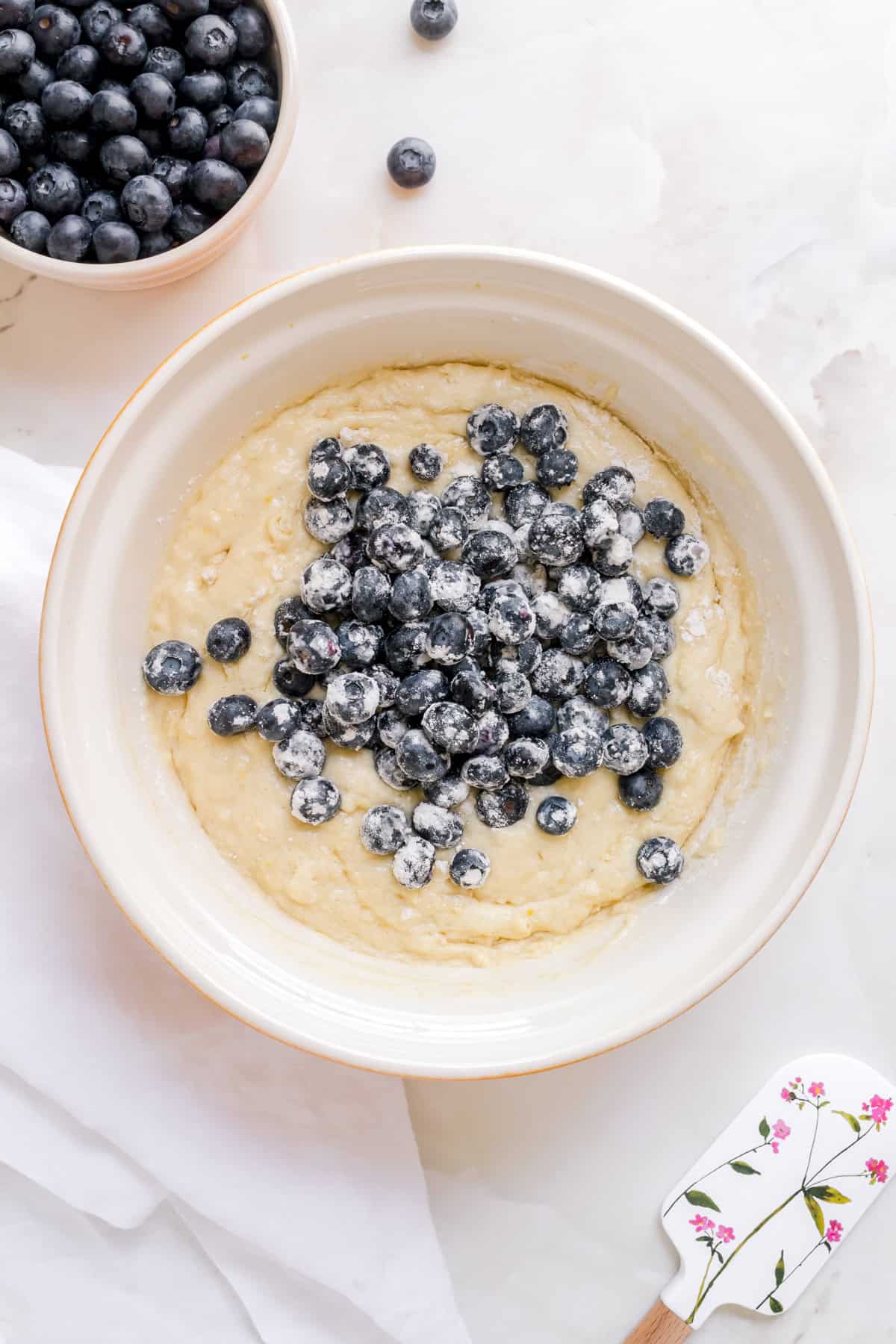 adding blueberries to discard muffin batter in ceramic bowl with spatula and bowl of blueberries.