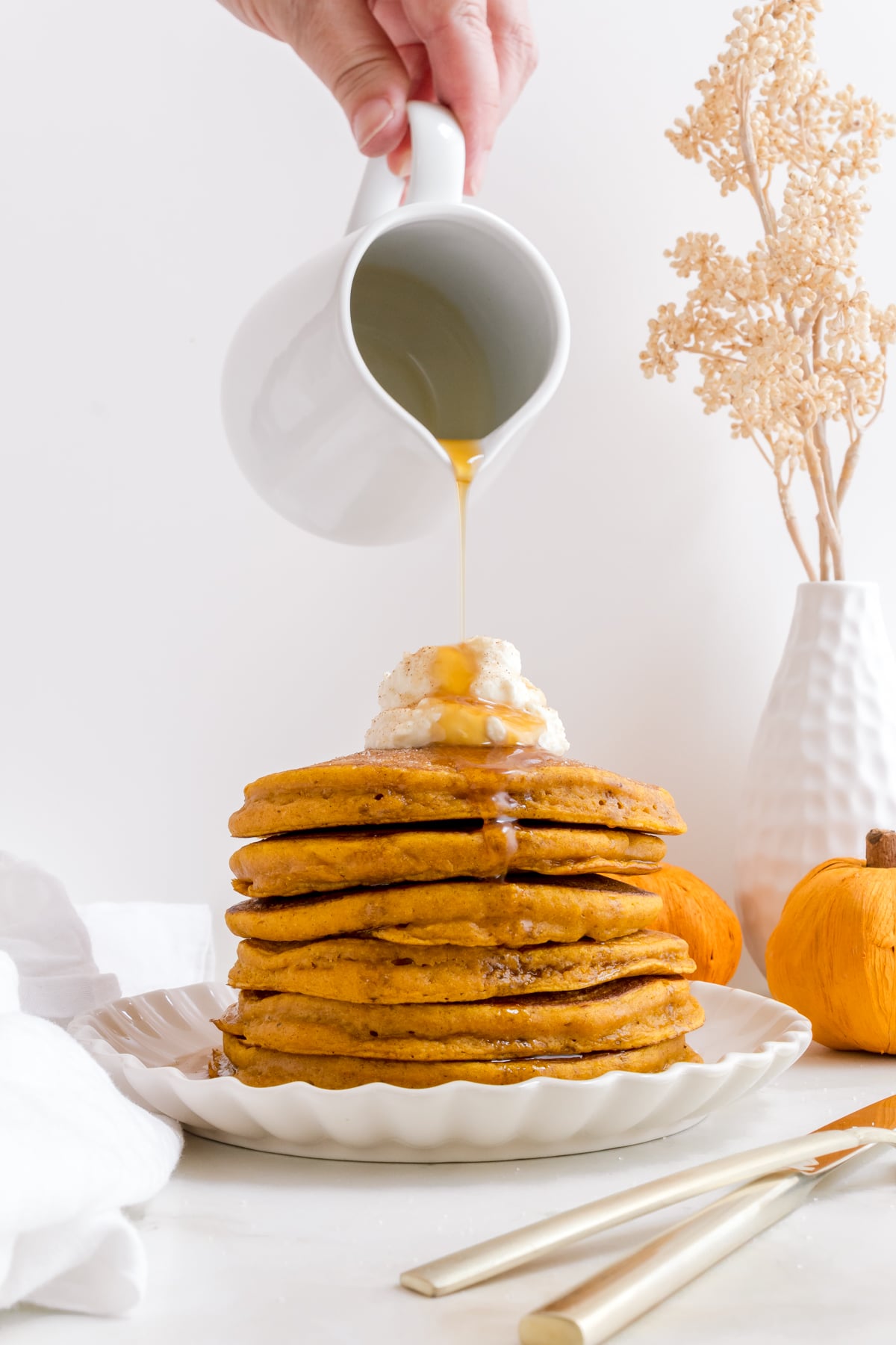 pouring maple syrup over a stack of pumpkin pancakes with whipped cream.