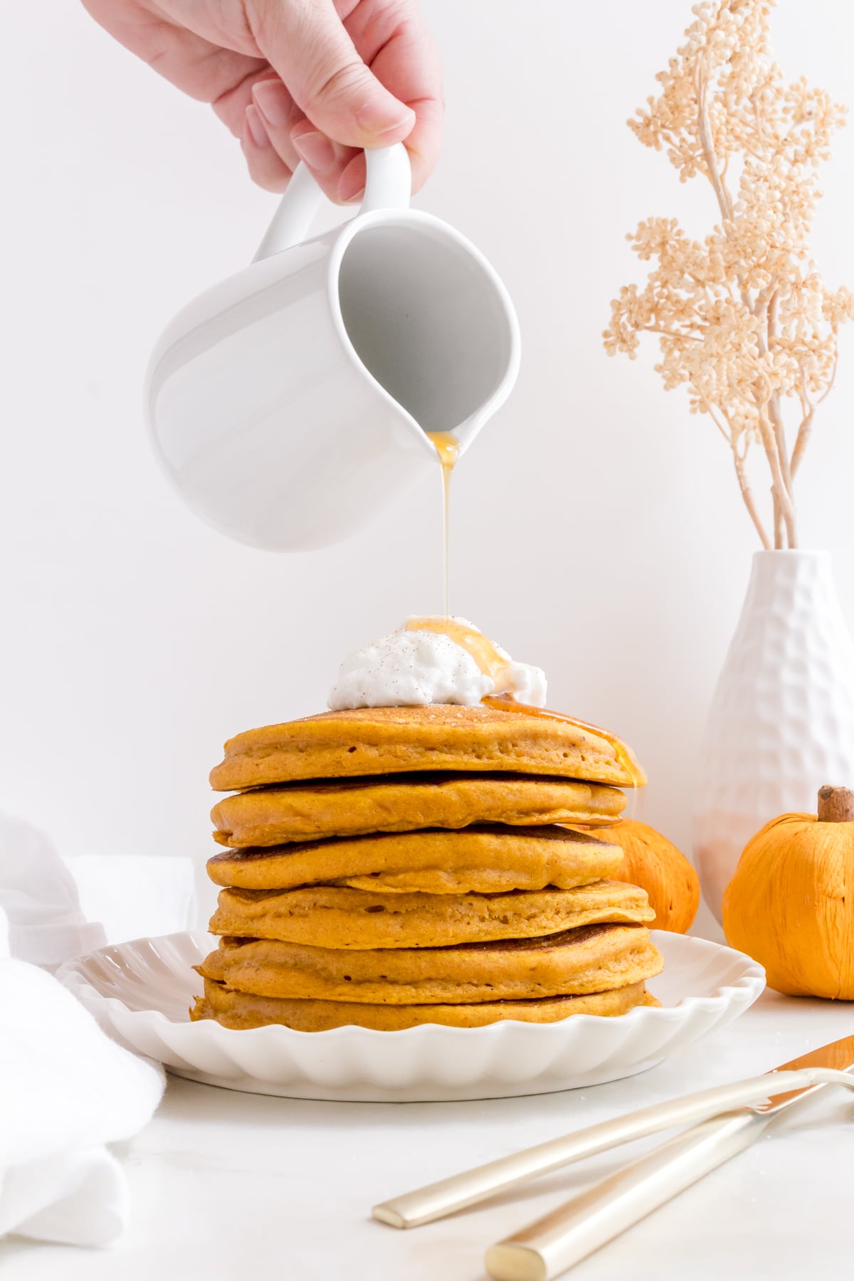 pouring syrup over a stack of pumpkin spice pancakes with whipped cream.