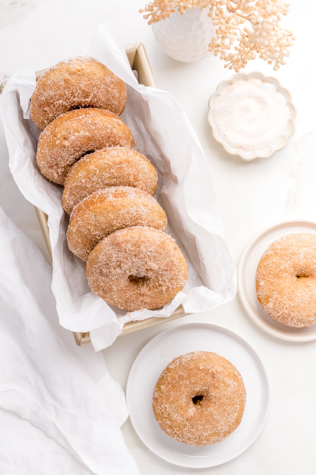 a gold bread pan of apple cider donuts and two round plates of donuts on a marble background with apple cider.