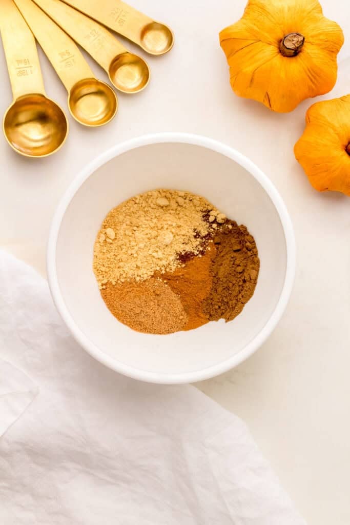 whisking spices for pumpkin pie spice recipe in a white mixing bowl with gold measuring cups.