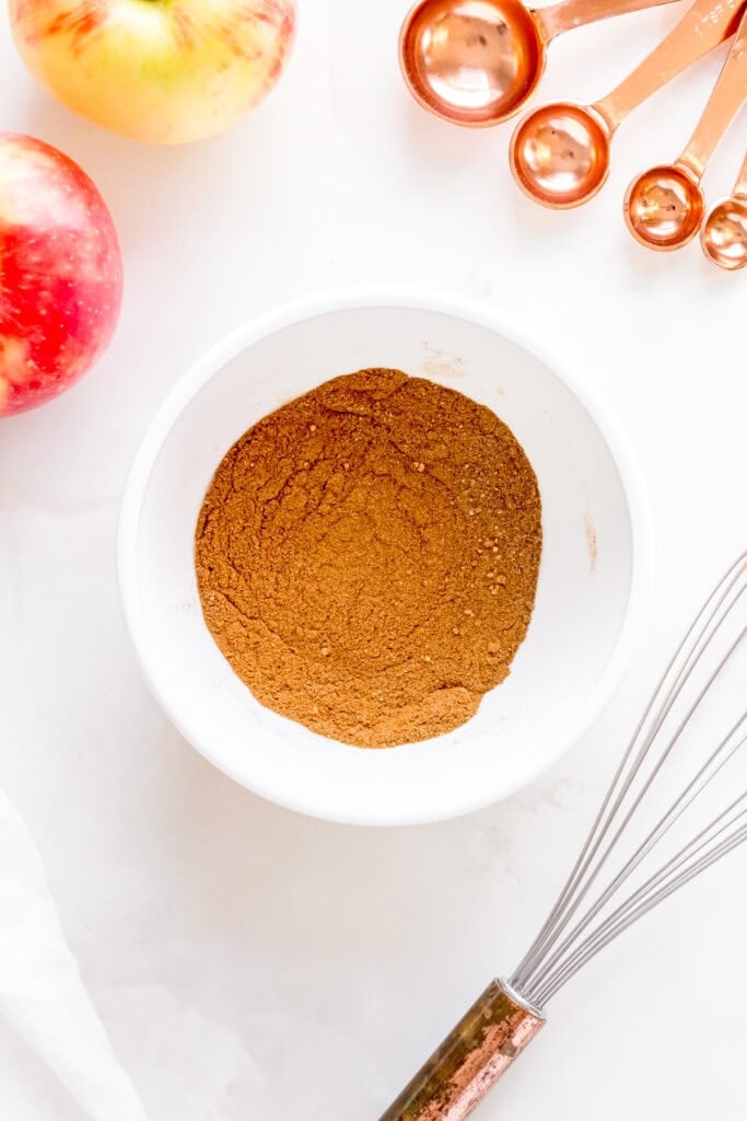apple pie spice in white mixing bowl with whisk and apples.