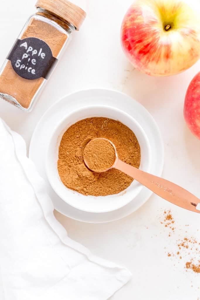 a bowl and jar of apple pie spice with a copper measuring spoon and apples.