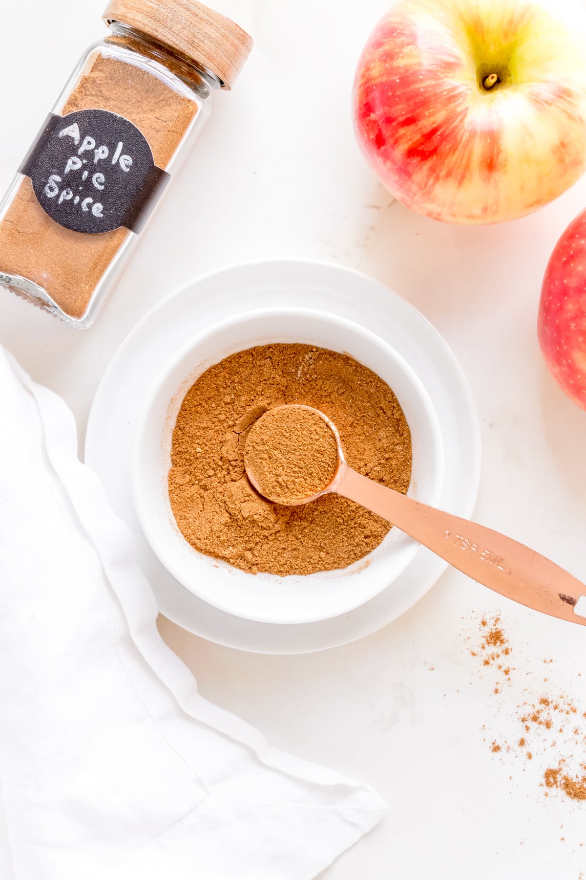 a bowl and jar of apple pie spice with a copper measuring spoon and apples.